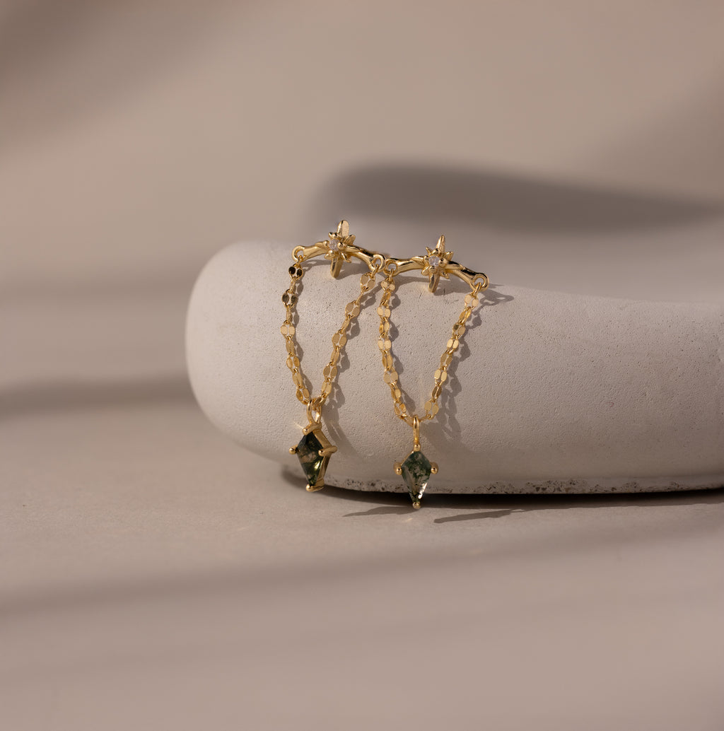 Agate Star Chain Earrings in 18K Gold feature gold chain drops with star-shaped studs and green agate gemstones, elegantly displayed on a smooth white stone.