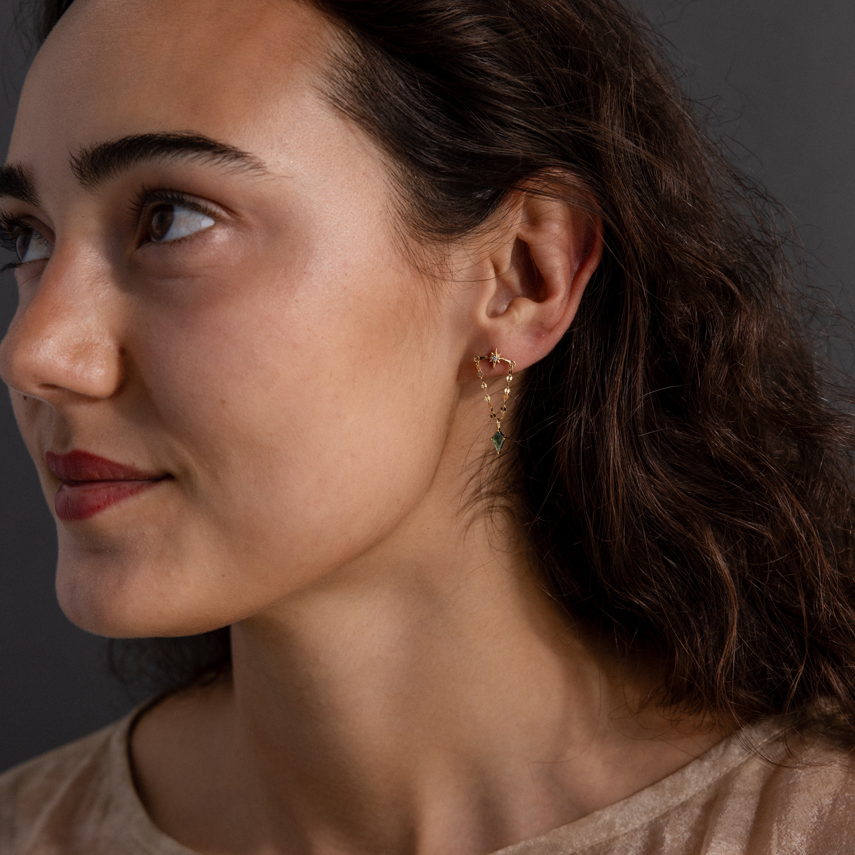 Woman with wavy brown hair wears Agate Star Chain Earrings in 18K Gold featuring green agate gemstones, looking to the left.