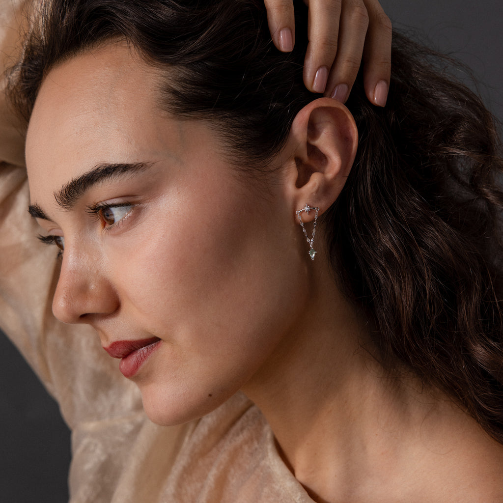 A woman with wavy brown hair wears Agate Star Chain Earrings in Sterling Silver, gazing to the side with her hand resting on her head.