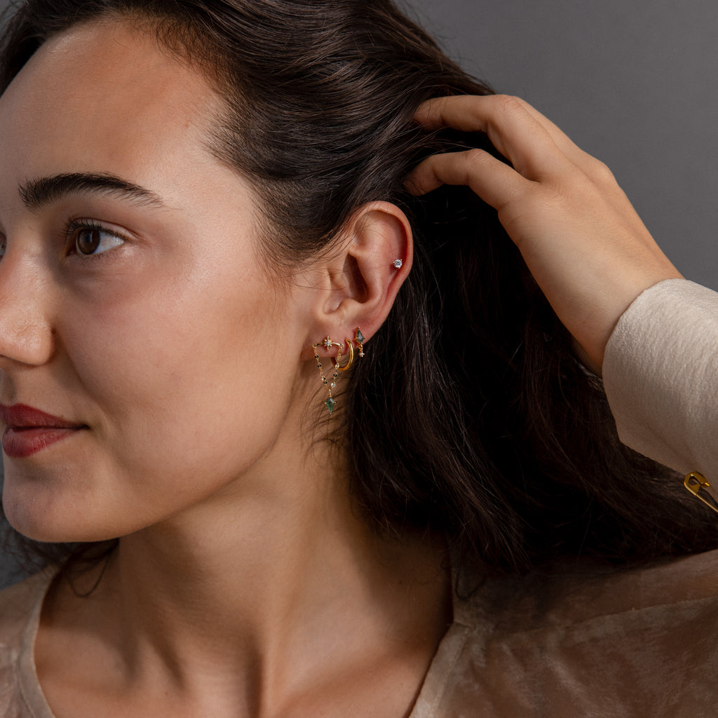 A woman with dark hair tucks her hair behind her ear, showcasing multiple colorful earrings, including the eye-catching Agate Star Chain Earrings in 18K Gold, featuring green agate for a playful celestial accent.