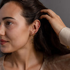 A dark-haired woman tucks her hair behind her ear, showcasing multiple gold earrings, including the Agate Star Chain Earrings, with green agate accents, set against a neutral background.