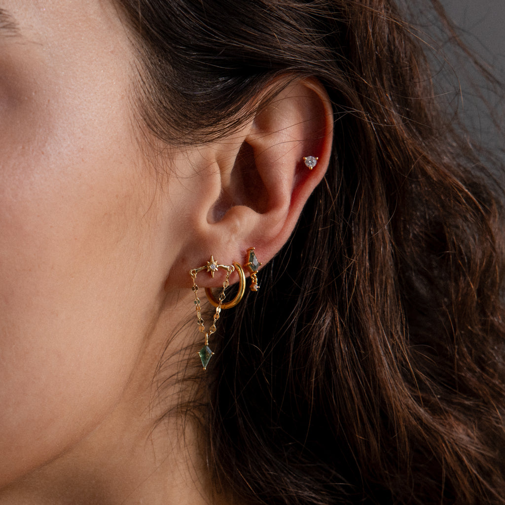 Close-up of a woman's ear adorned with multiple gold earrings, featuring the Agate Star Chain Earrings in 18K Gold and a small stud in the upper cartilage for a chic celestial look.