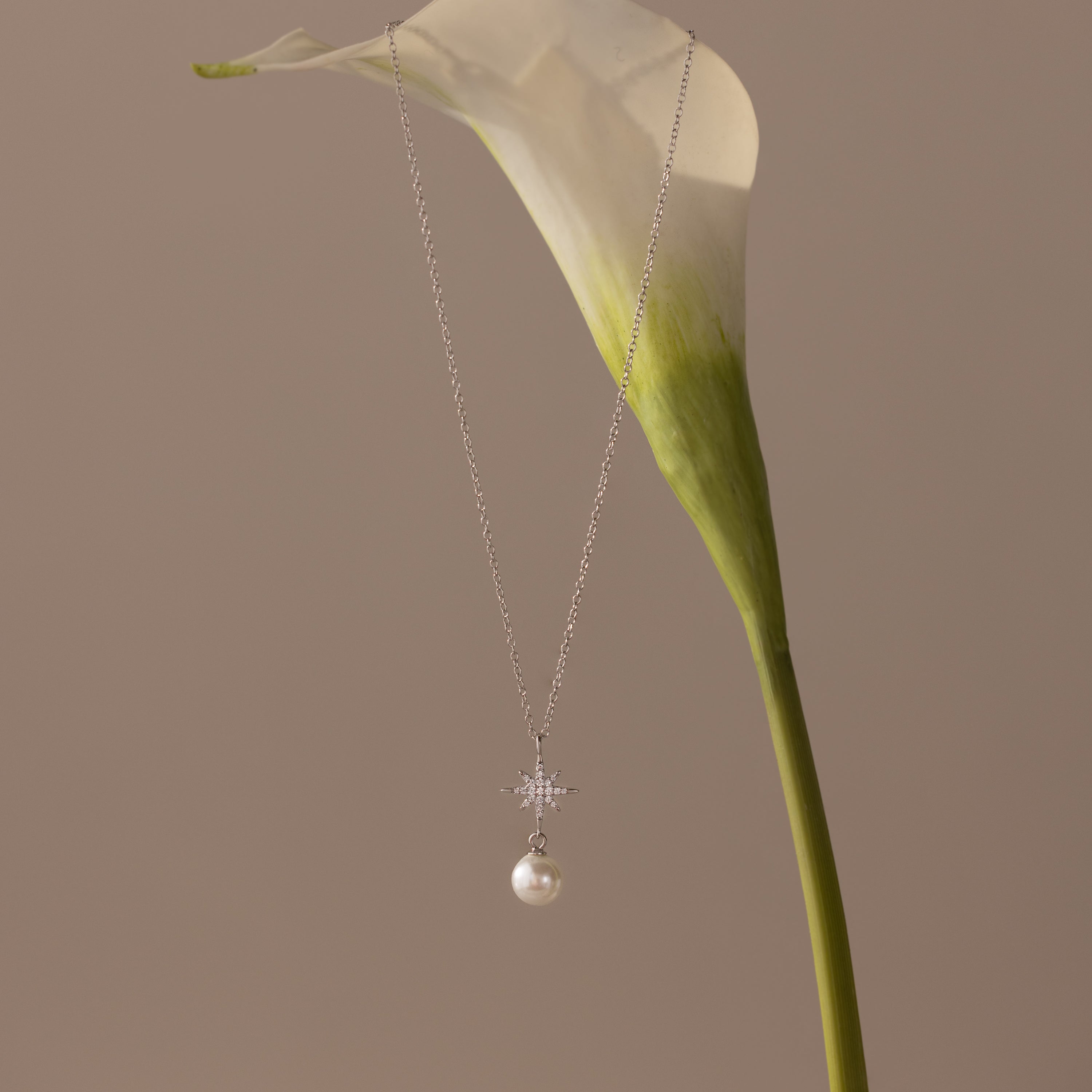 The North Star Pearl Necklace in Sterling Silver, featuring a pearl pendant, is displayed on a white calla lily with a beige background.