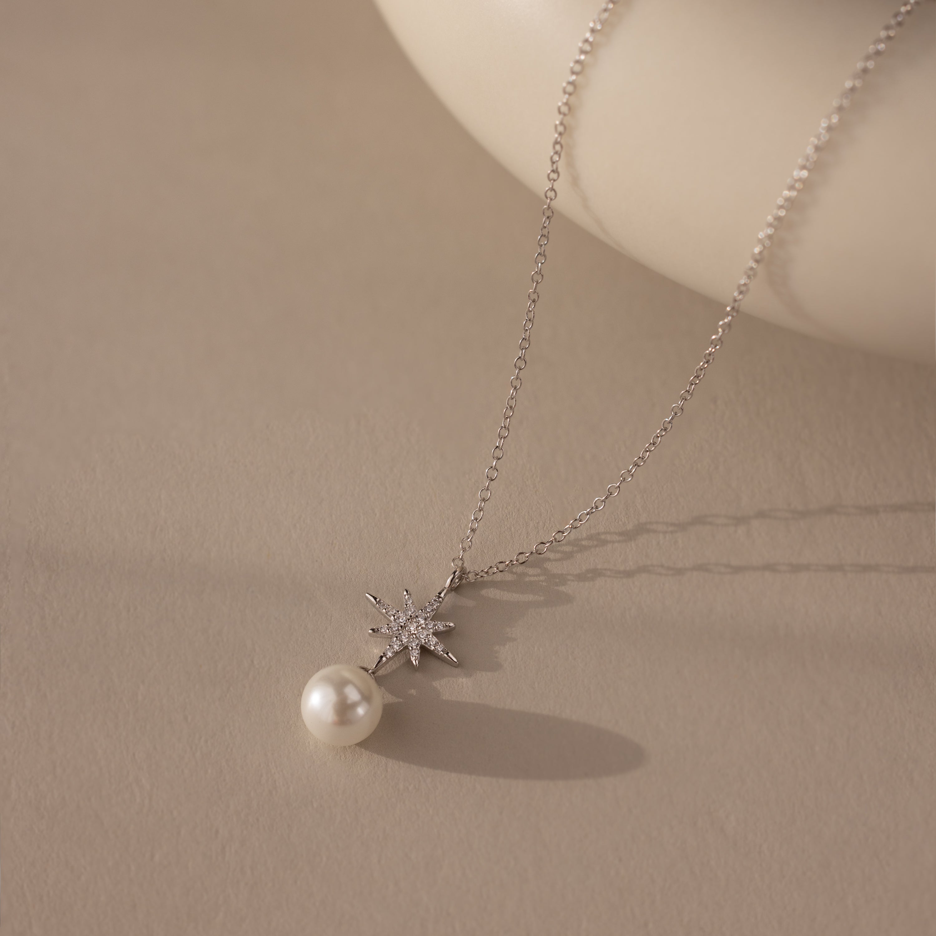 The North Star Pearl Necklace in Sterling Silver features a star-shaped pendant with a dangling pearl, elegantly displayed on a neutral surface.