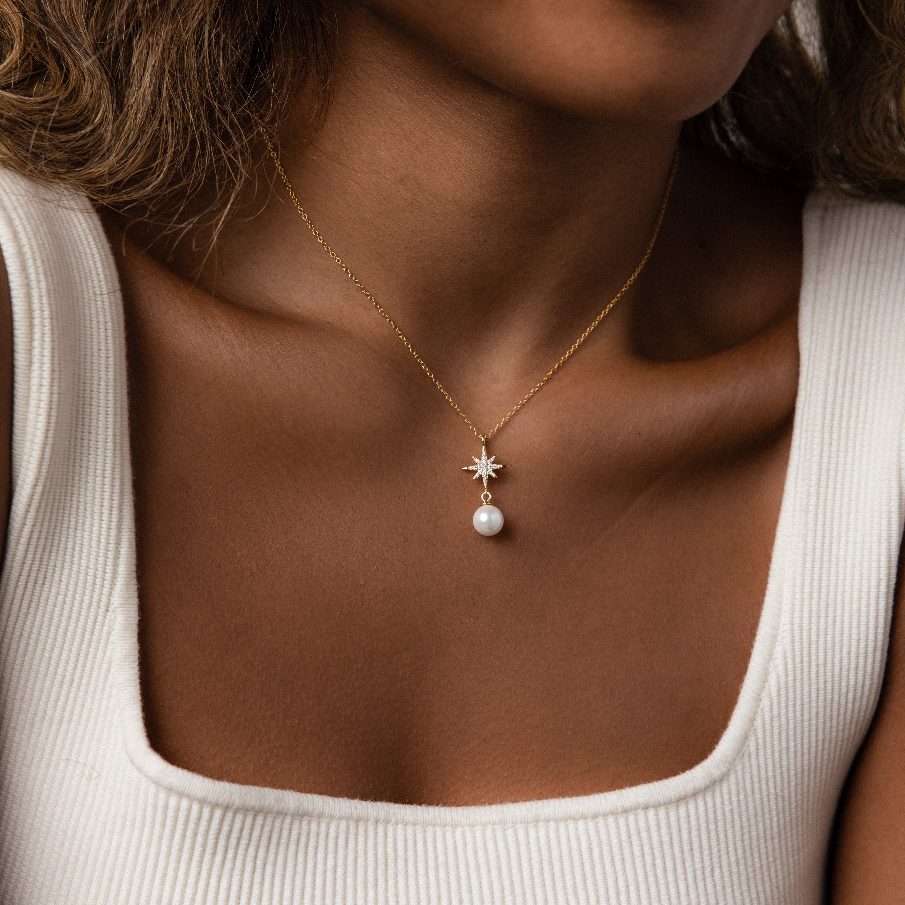 A woman in a white square-neck top wears the North Star Pearl Necklace, featuring a gold chain with star and pearl pendant.