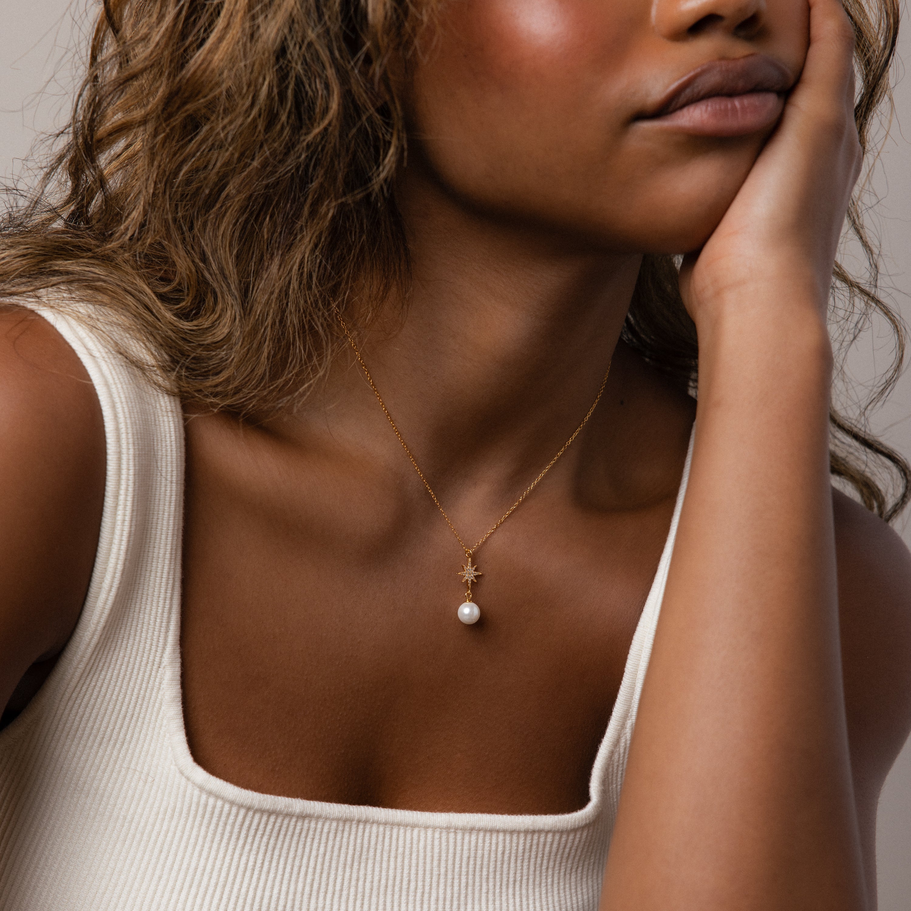 A woman in a white tank top wears the North Star Pearl Necklace in 18K Gold, its star pendant and lustrous pearl adding elegance as she rests her chin on her hand.