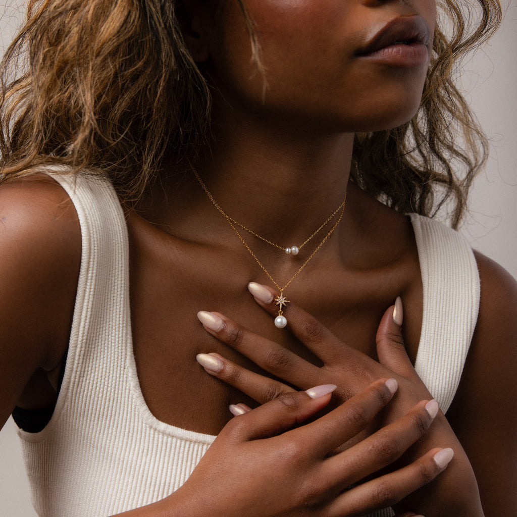 A woman in a white tank top layers gold necklaces, highlighting the North Star Pearl Necklace in 18K Gold as she gently touches her chest to showcase the elegant pearl jewelry.