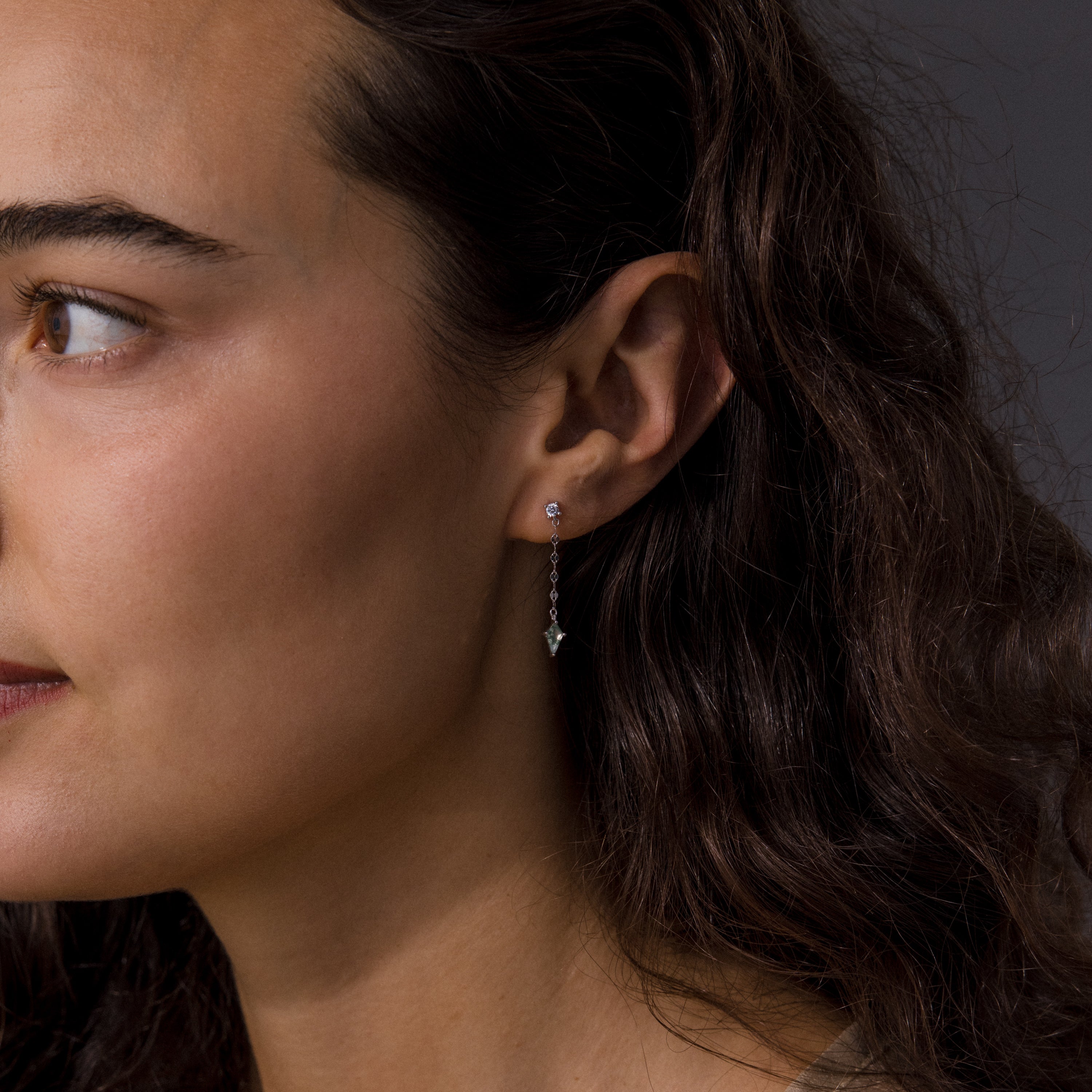 A woman with wavy brown hair wears Agate Diamond Dangle Earrings in Sterling Silver, shown in a close-up side profile to highlight their shimmering detail.