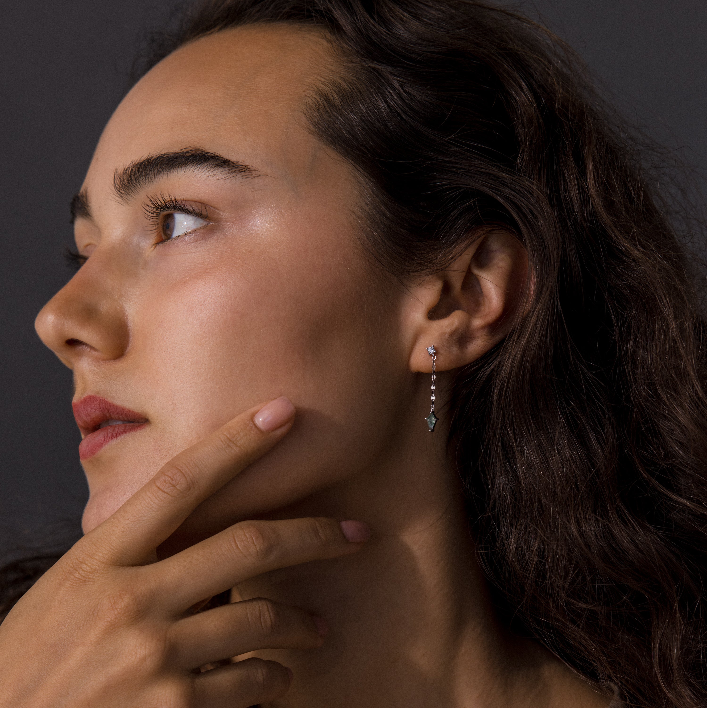 Woman with long dark hair looks left, touching her face, wearing Agate Diamond Dangle Earrings in Sterling Silver against a dark background.