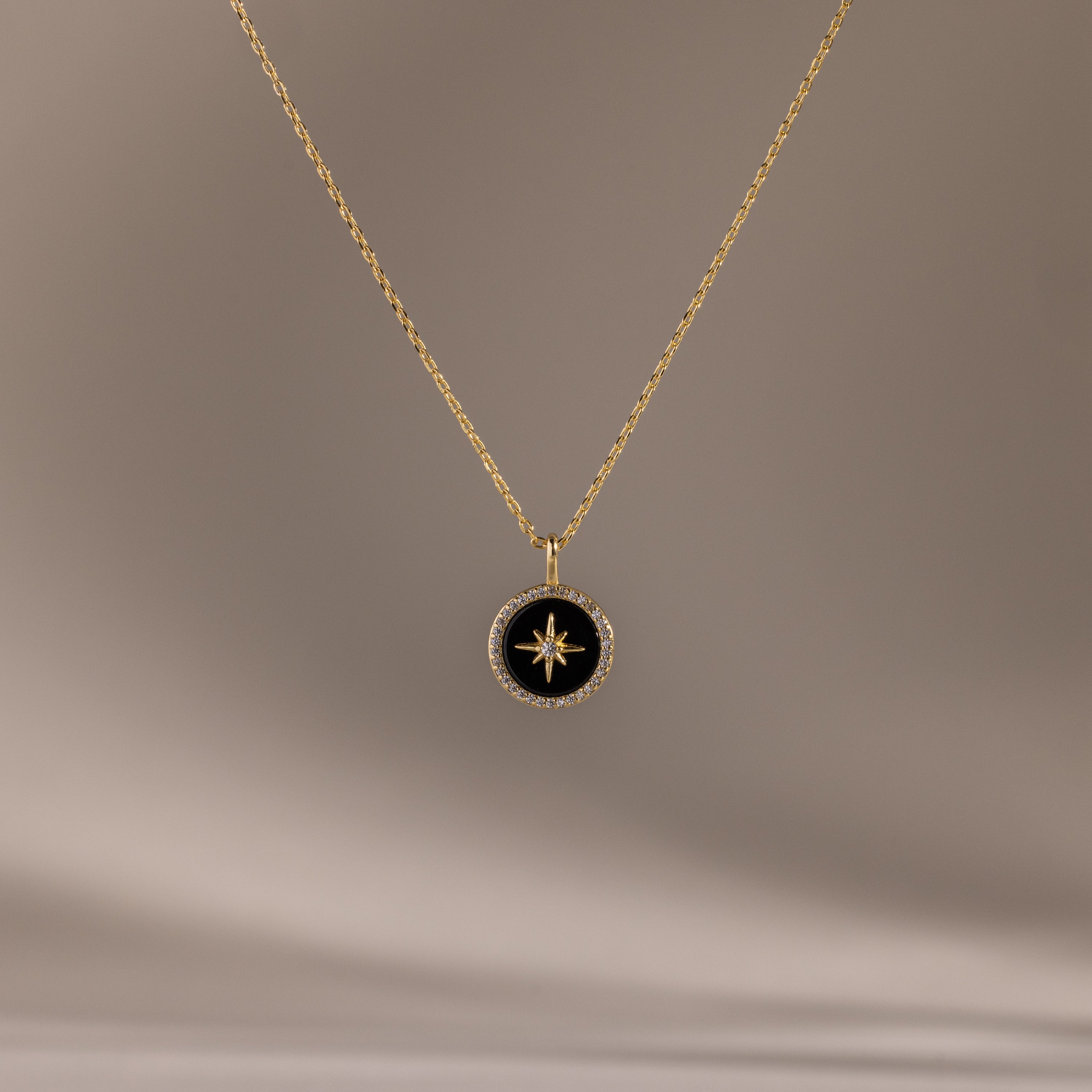 The Onyx Star Pendant Necklace showcases a round black onyx pendant with a central star design and a diamond-like border, making this celestial accessory shine beautifully against any soft background.