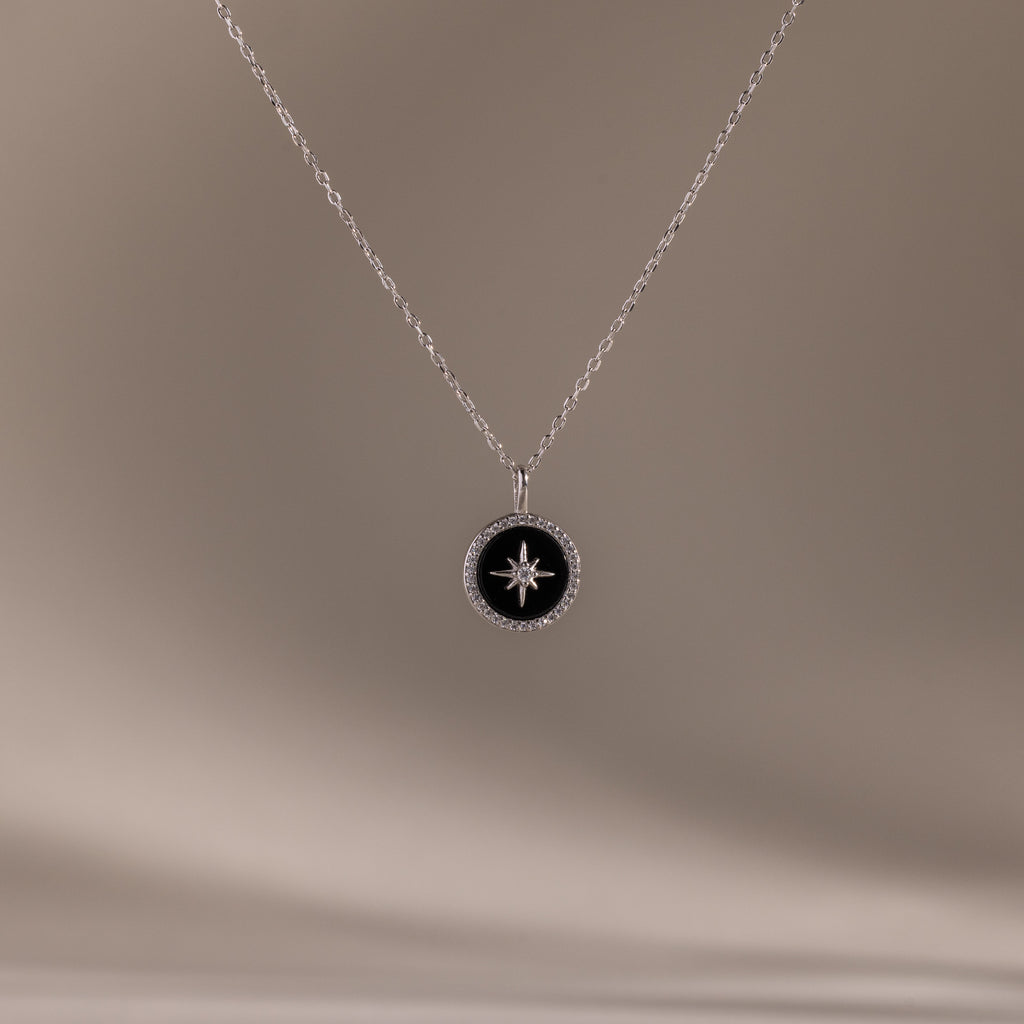 The Onyx Star Pendant Necklace in Sterling Silver features a round black onyx with a celestial star design, shown against a soft beige background.