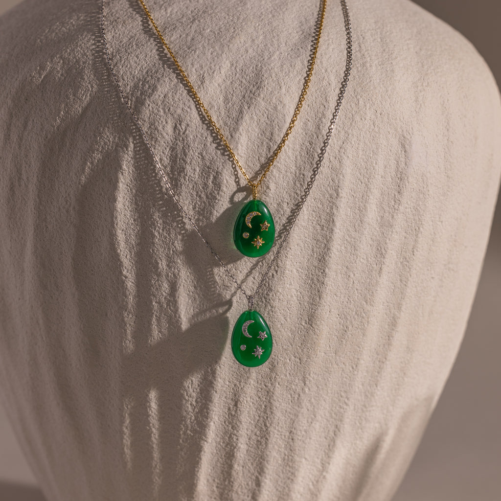 The Celestial Jade Pendant Necklace features two jade pendants on gold and silver chains, each with star and moon accents, creating an elegant celestial look. The necklace is showcased on a white bust.