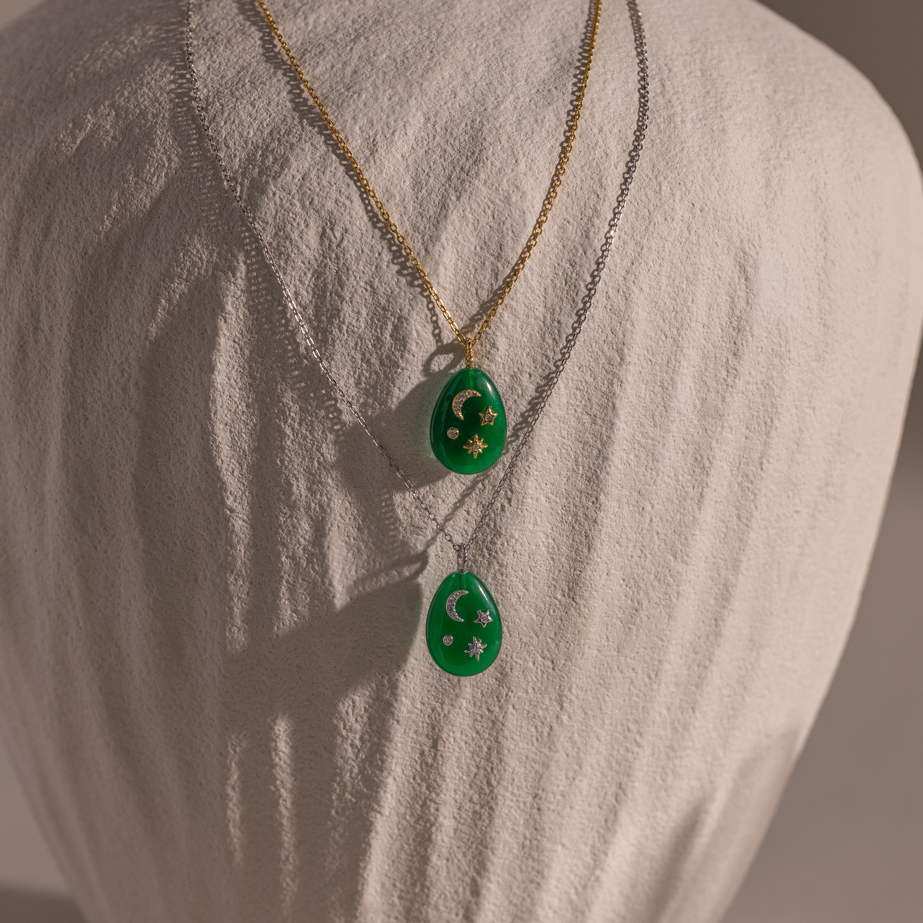 The Celestial Jade Pendant Necklace features two jade pendants on gold and silver chains, each with star and moon accents, creating an elegant celestial look. The necklace is showcased on a white bust.