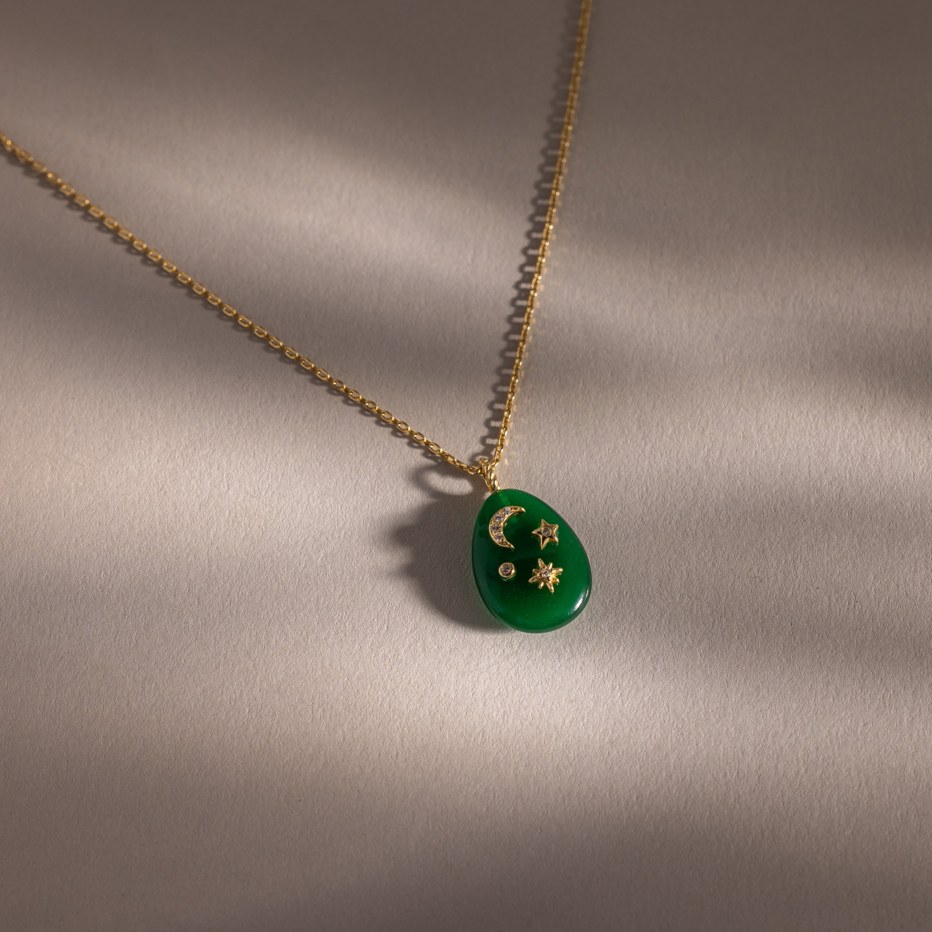 The Celestial Jade Pendant Necklace features a gold chain with a green teardrop gemstone and celestial moon, star, and sun motifs, shown on a neutral background.