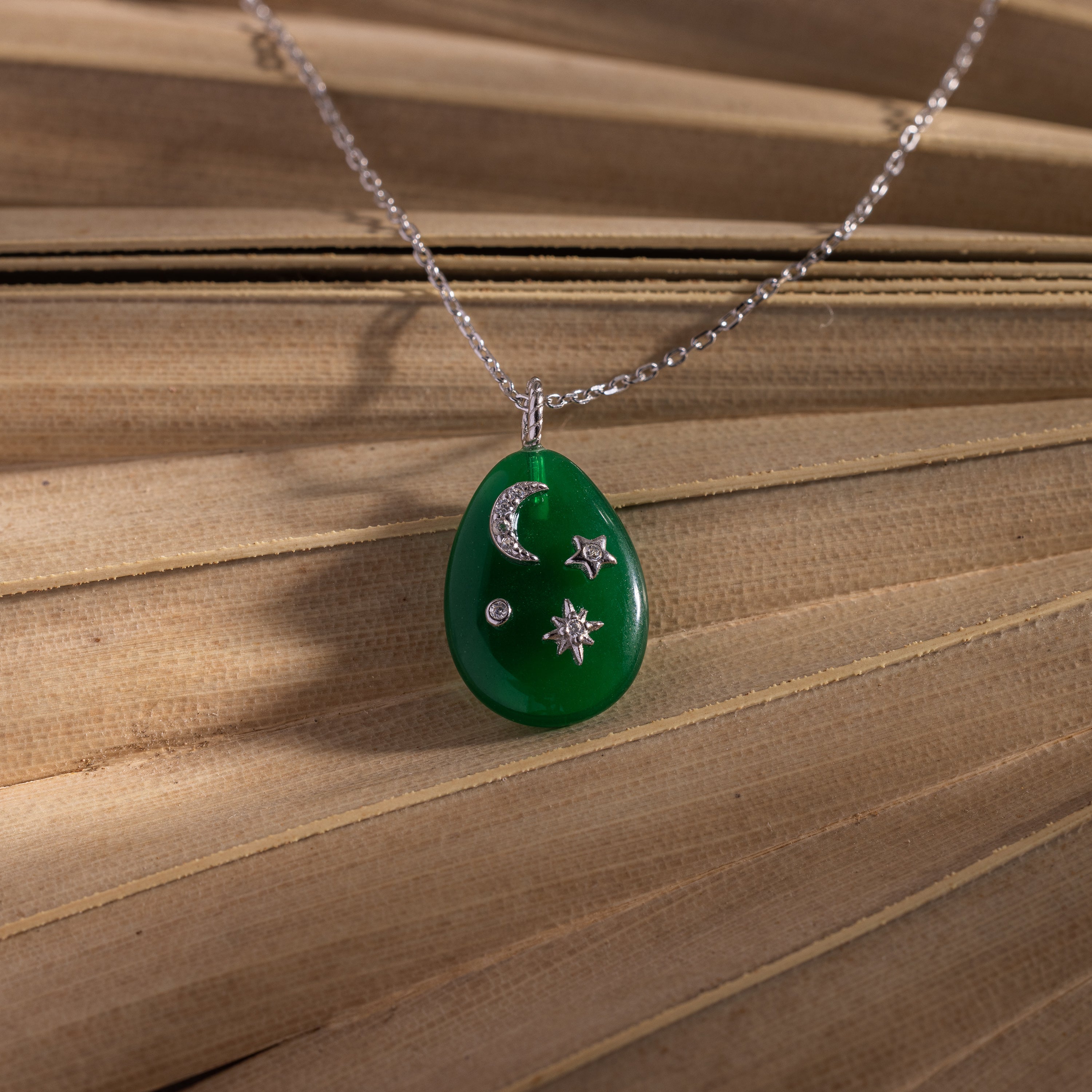 The Celestial Jade Pendant Necklace, featuring moon, star, and gem designs, hangs on a silver chain against a wooden background.