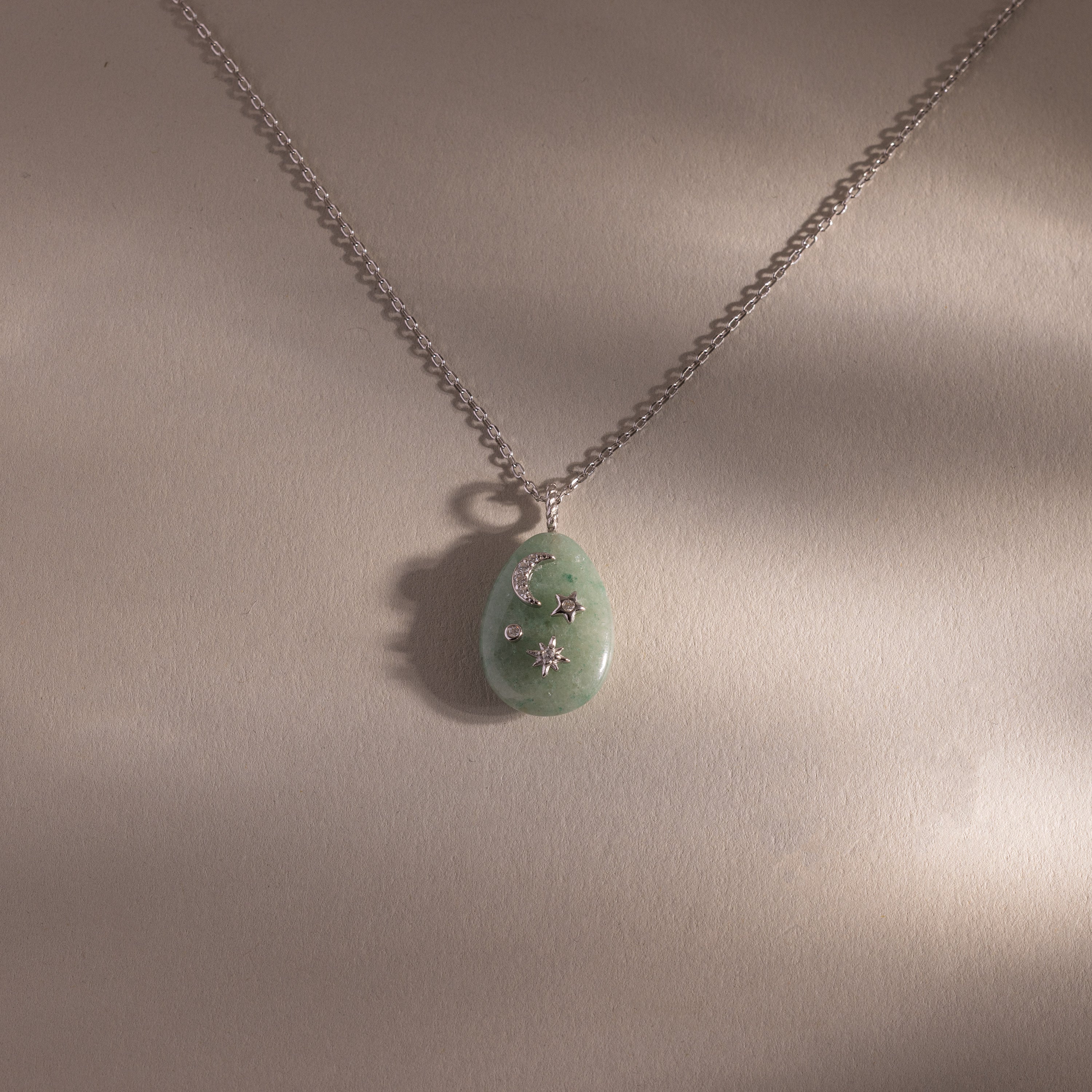 The Celestial Aventurine Pendant Necklace features a green aventurine pendant with intricate silver crescent moon and star accents on a beige background.