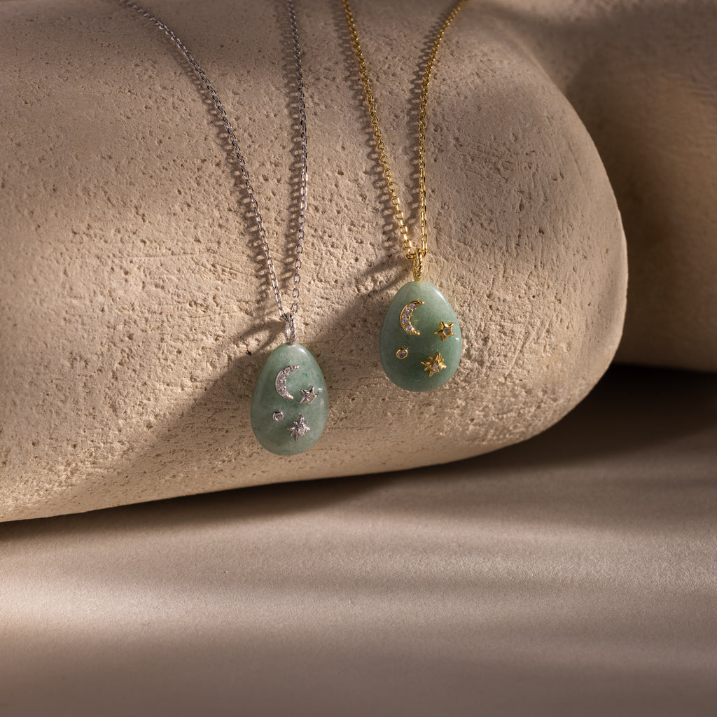 The Celestial Aventurine Pendant Necklace features crescent moon and star charms with green aventurine accents on silver and gold chains, beautifully displayed on textured stone.