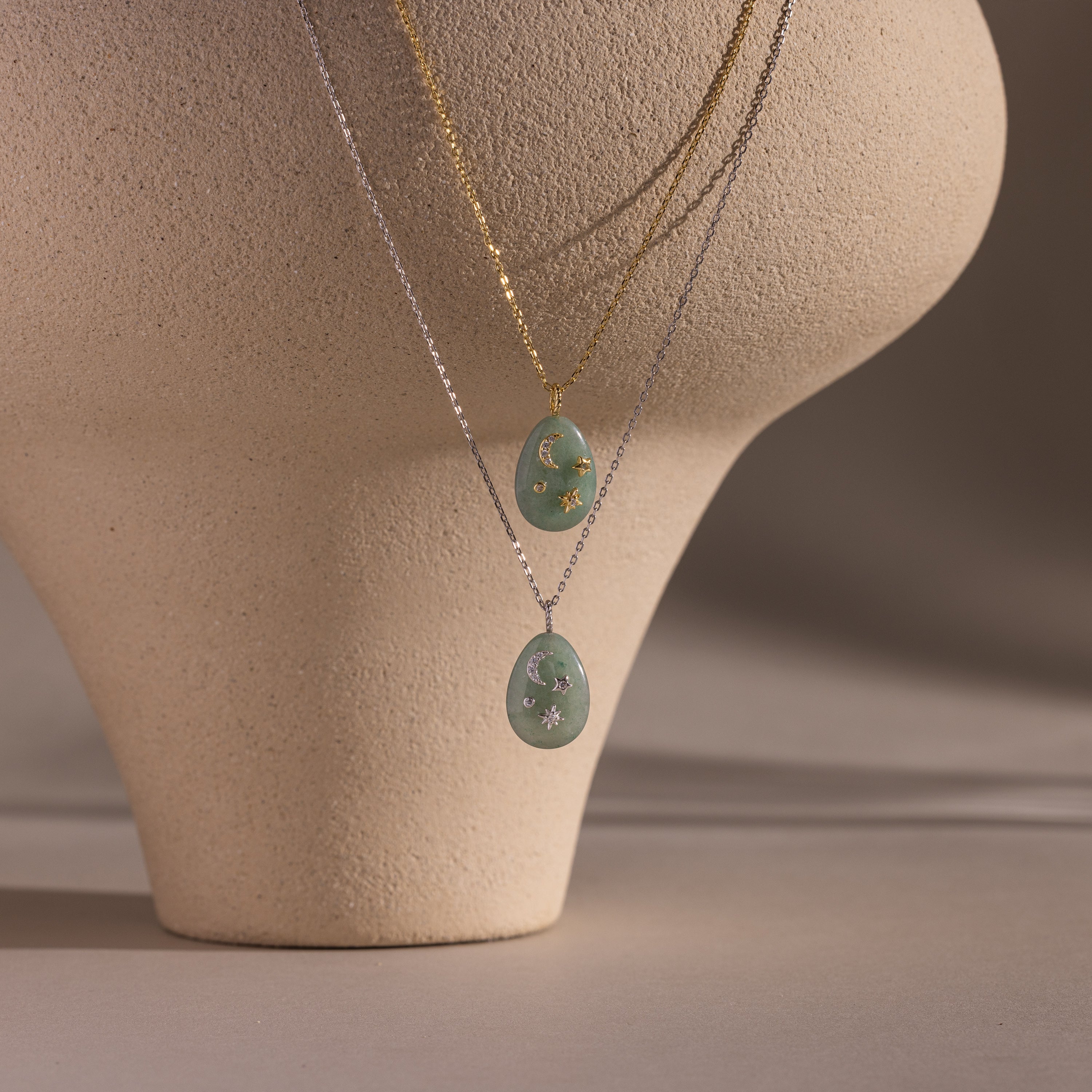 Two layered necklaces with oval green gemstone pendants, including the Celestial Aventurine Pendant Necklace, displayed on a textured beige mannequin.