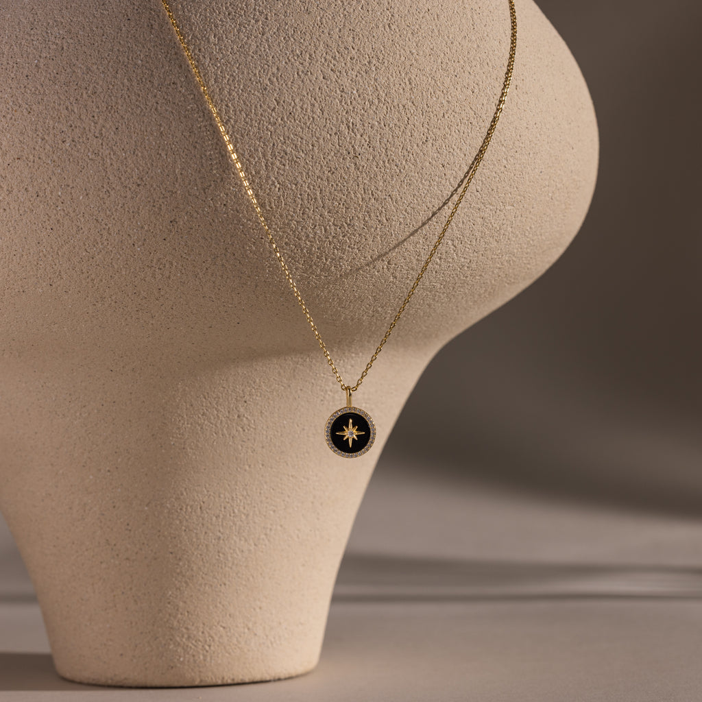 The Onyx Star Pendant Necklace in 18K Gold features a round black onyx pendant with a star design, beautifully displayed on a textured, cream-colored sculpture.