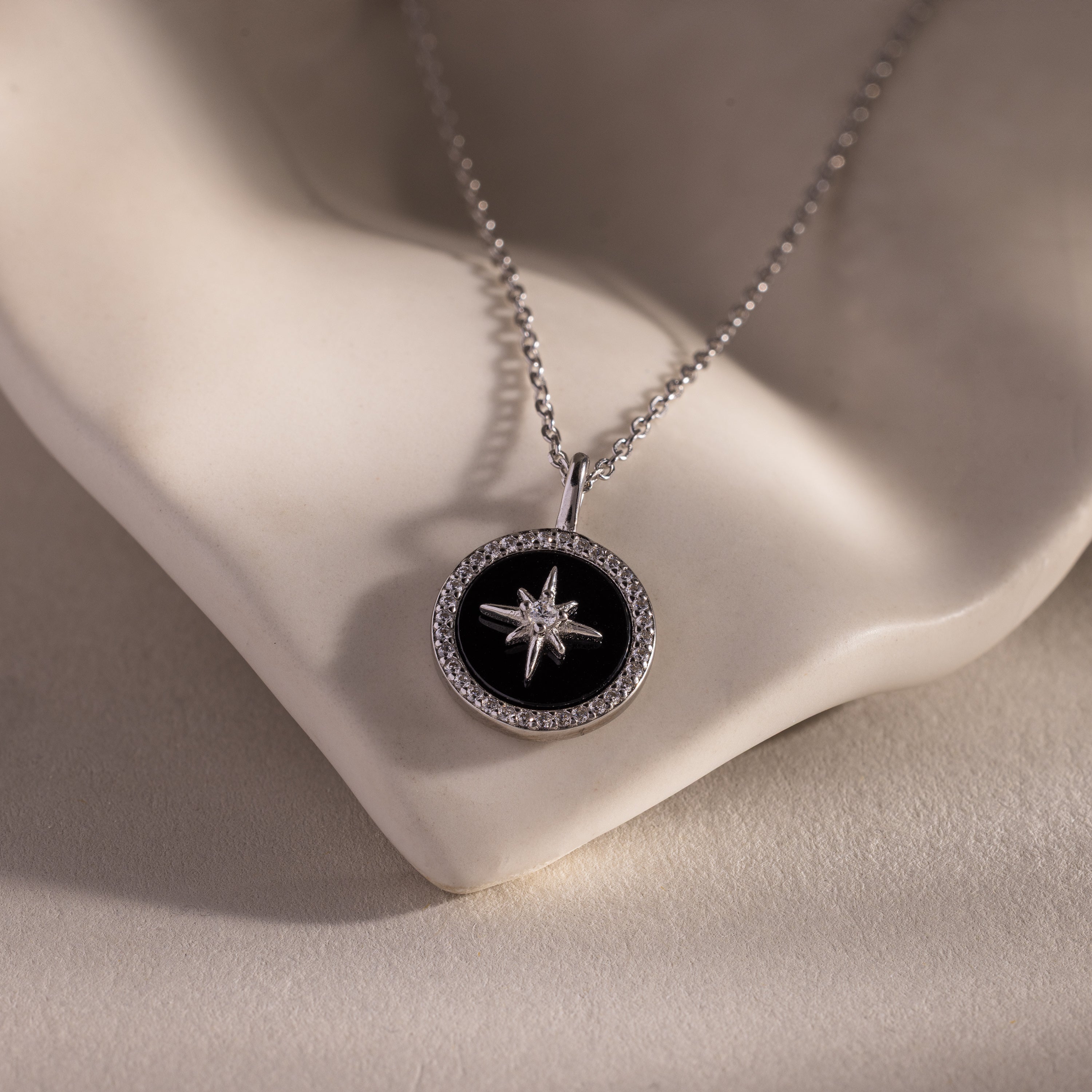 The Onyx Star Pendant Necklace features a round black onyx star pendant on a silver chain, beautifully displayed on a white sculpted surface—perfect for adding celestial elegance to any outfit.