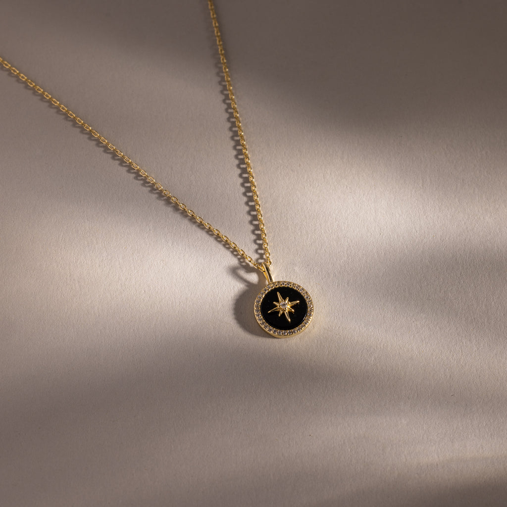 Onyx Star Pendant Necklace—a gold necklace with a black onyx circular pendant featuring a star design, displayed on a light beige surface. A striking celestial accessory for any occasion.