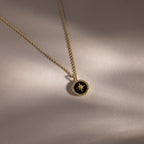 Onyx Star Pendant Necklace—a gold necklace with a black onyx circular pendant featuring a star design, displayed on a light beige surface. A striking celestial accessory for any occasion.