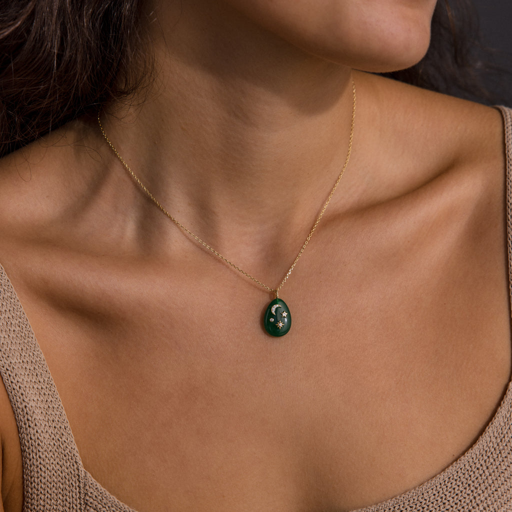 A woman wears the Celestial Jade Pendant Necklace, featuring a gold chain and a green oval charm, shown against her bare neck and collarbone.