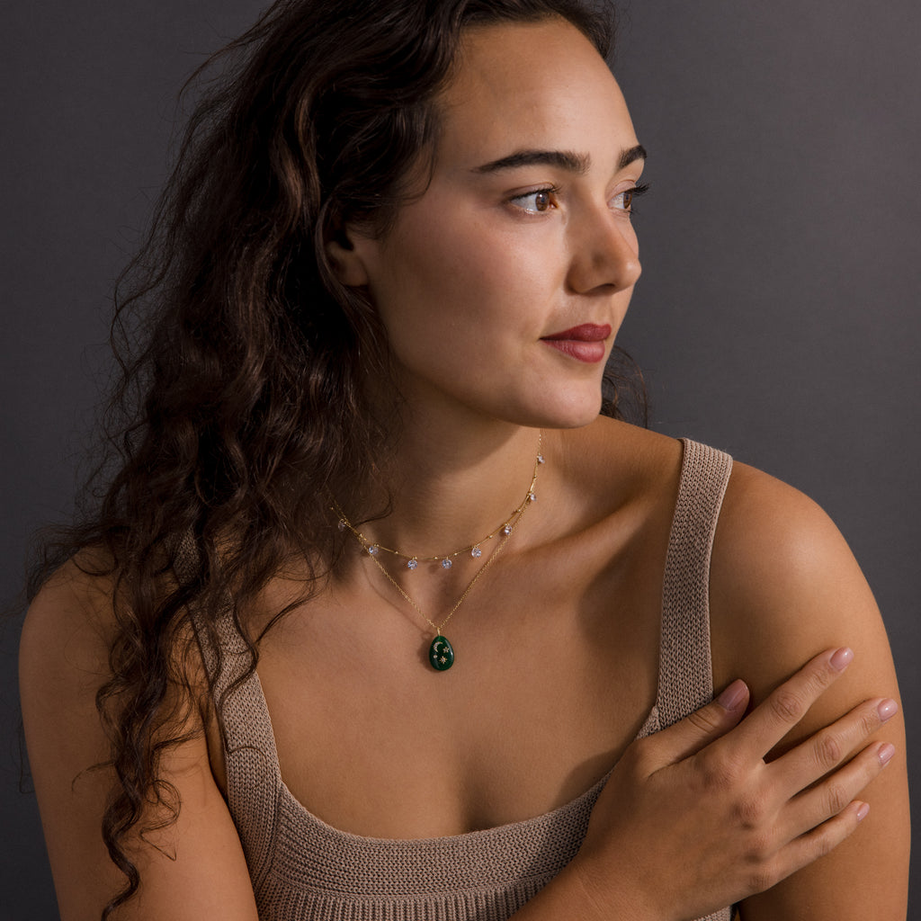 A woman with long curly hair in a beige top wears the Celestial Jade Pendant Necklace, layered with other pieces, as she looks to the side against a gray background.