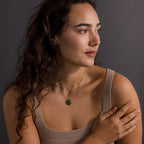 A woman with long curly hair in a beige top wears the Celestial Jade Pendant Necklace, layered with other pieces, as she looks to the side against a gray background.