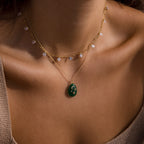 Close-up of a woman's neck wearing two delicate gold necklaces—a green gemstone necklace and the Celestial Jade Pendant Necklace—against tan skin.