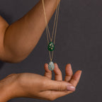 A hand holds two Celestial Gemstone Pendant Necklaces, each featuring green gemstones and star accents, beautifully showcasing the magic of Moon and Stars Jewelry against a dark background.