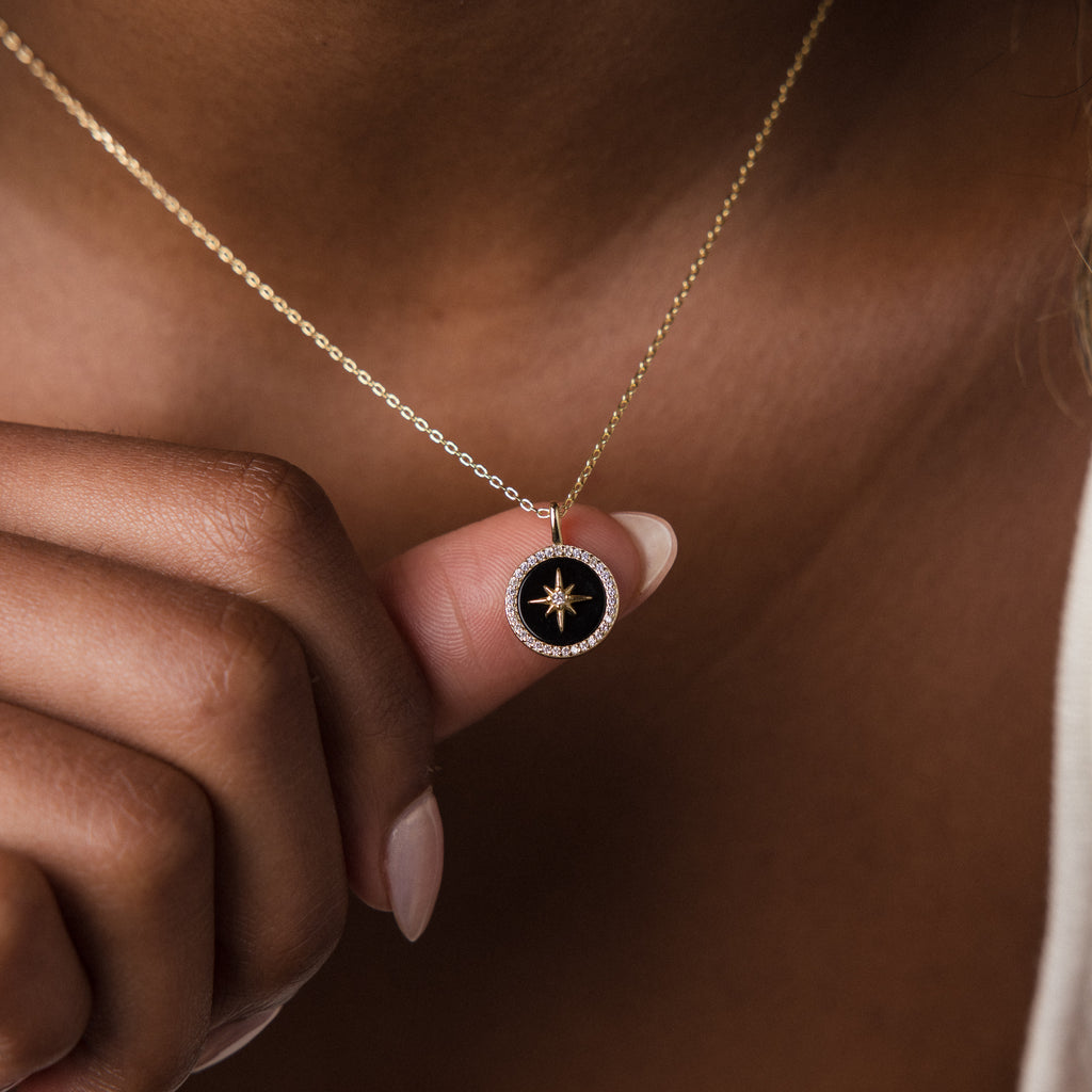 A hand holds the Onyx Star Pendant Necklace, featuring a round black onyx pendant adorned with a striking celestial star design.