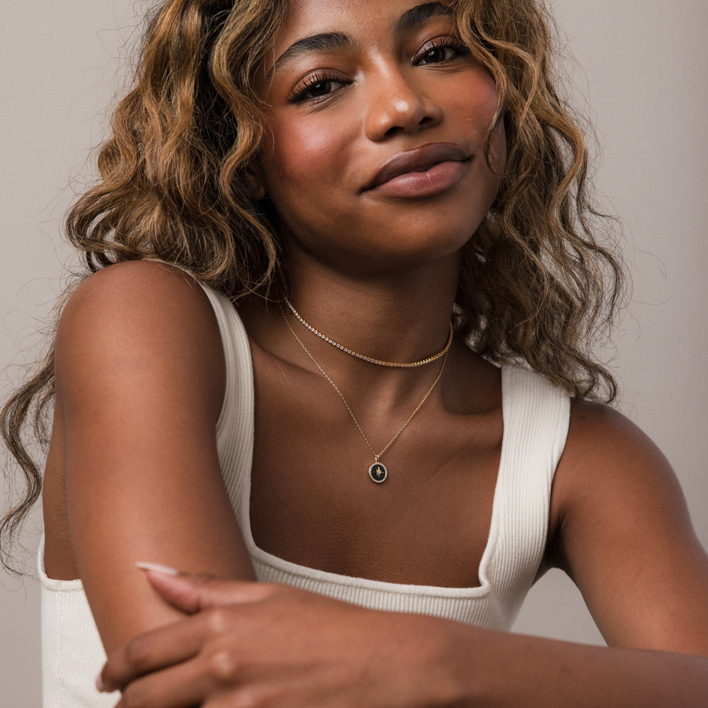 A woman with wavy hair in a white tank top smiles gently at the camera, wearing layered necklaces including the Onyx Star Pendant Necklace.