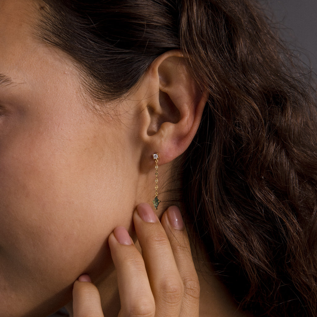 A person touches their ear, highlighting modern essentials: Agate Diamond Dangle Earrings featuring a gold chain, a green agate-like gem, and a clear stone stud.