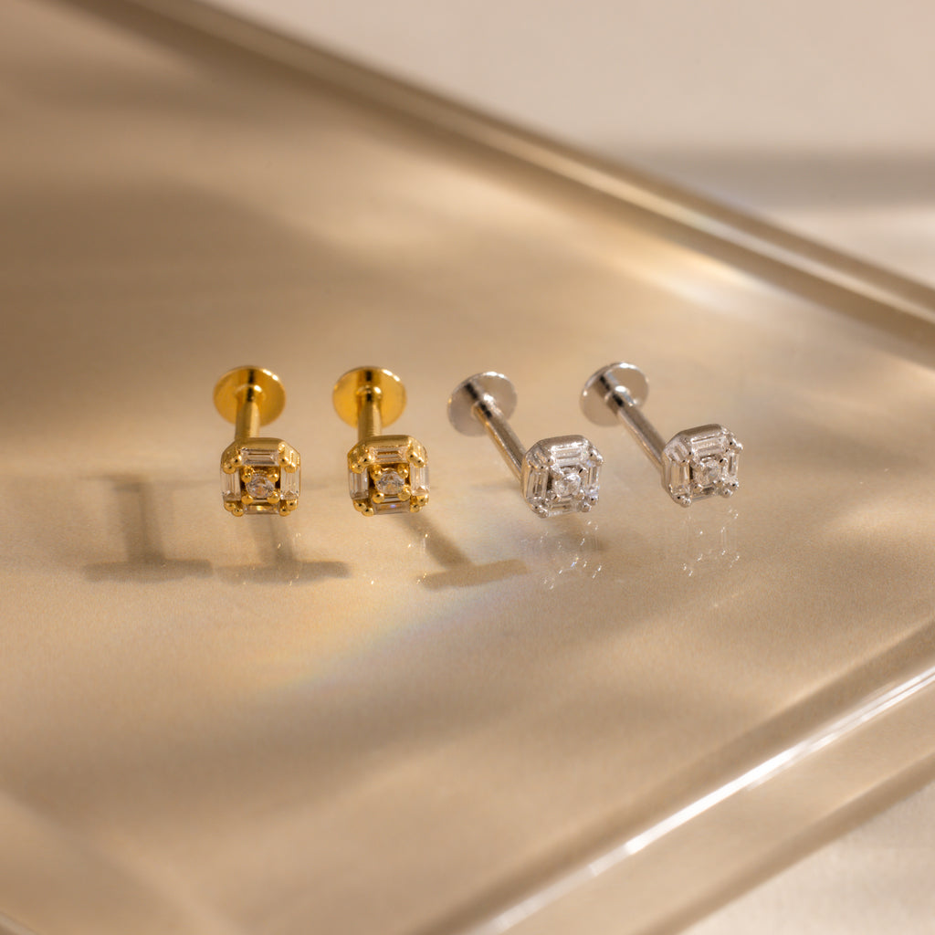Two pairs of Art Deco flatback stud earrings—one in gold and one in silver—featuring baguette-cut diamonds, displayed on a reflective, warm-toned surface.