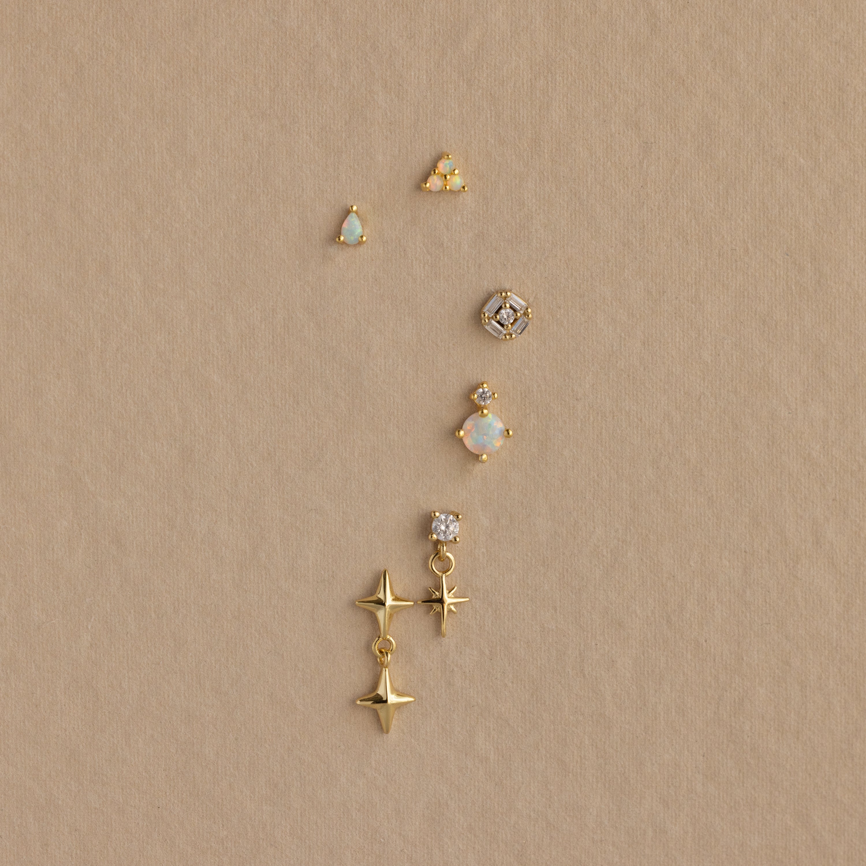 Six Opal  Diamond Flatback Studs in 18K Gold, featuring various designs like stars and gems, are arranged vertically on a beige background.