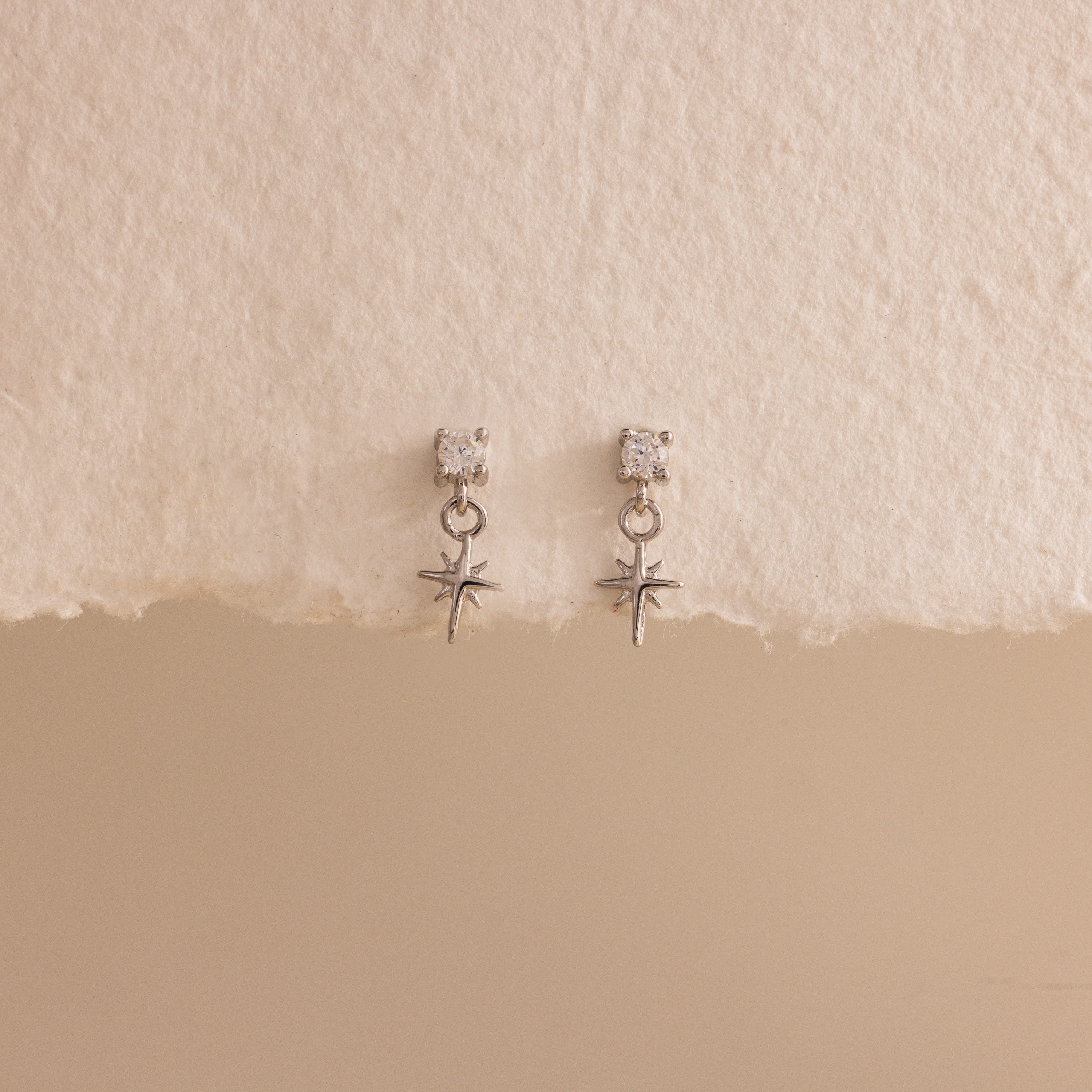 A straight-on product display of the 18k silver Diamond Star Flatback Earrings against a soft beige and off-white background, highlighting the charm's clean symmetry.