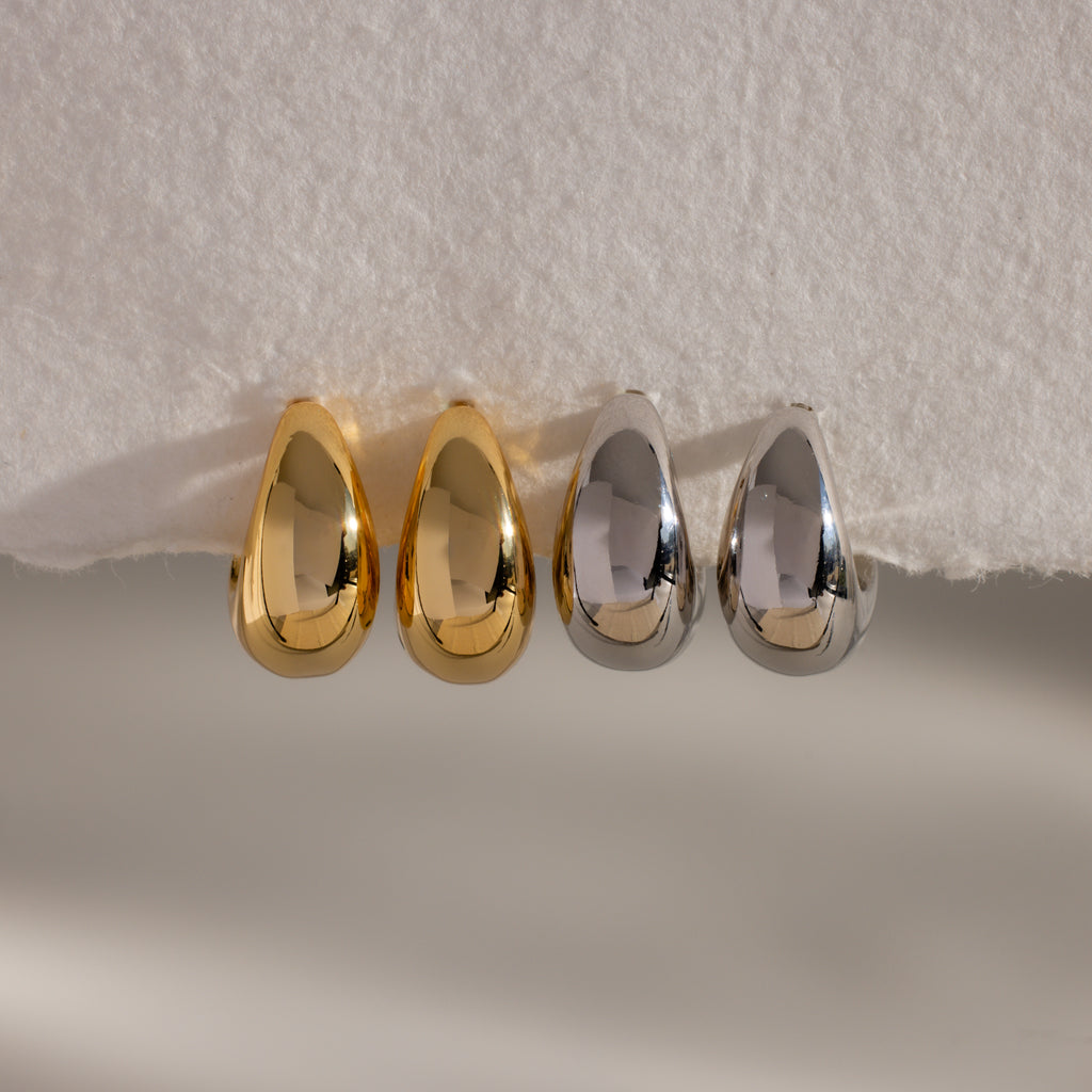 Four Bold Teardrop Earrings—two gold and two silver—are displayed on a white textured surface, highlighting modern jewelry with a striking flair.