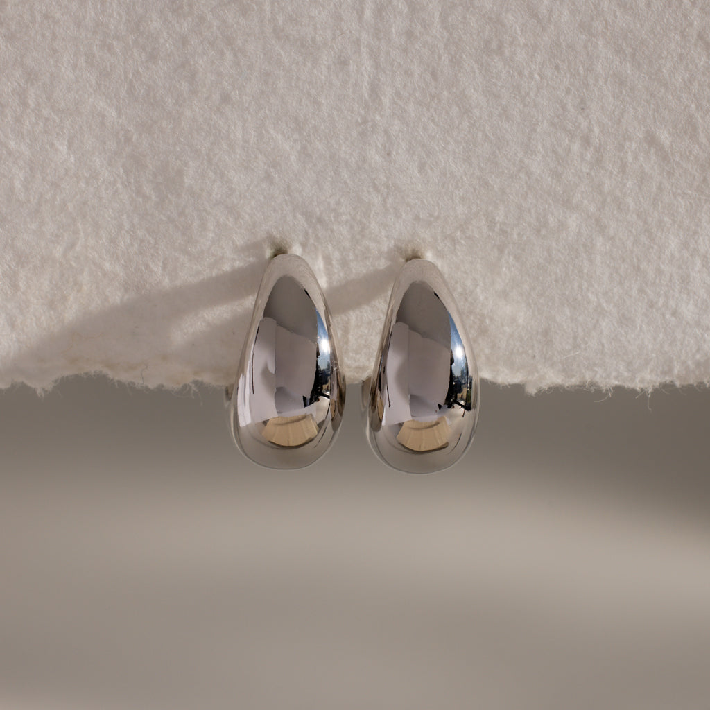 The Bold Teardrop Earrings in White Gold feature a striking teardrop silhouette and shiny finish, resting on a textured white surface—perfect for making a statement.