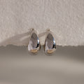The Bold Teardrop Earrings in White Gold feature a striking teardrop silhouette and shiny finish, resting on a textured white surface—perfect for making a statement.