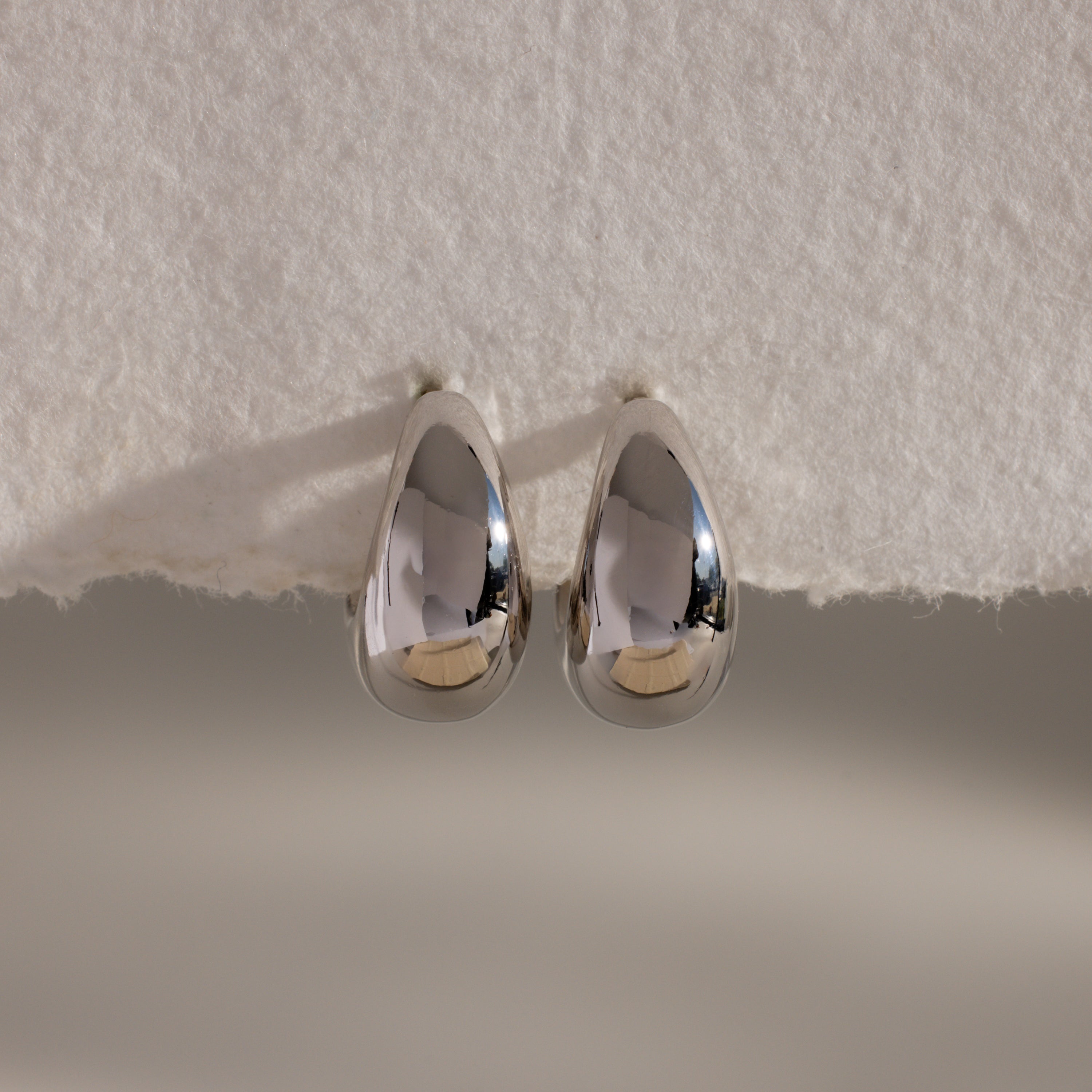 The Bold Teardrop Earrings in White Gold feature a striking teardrop silhouette and shiny finish, resting on a textured white surface—perfect for making a statement.
