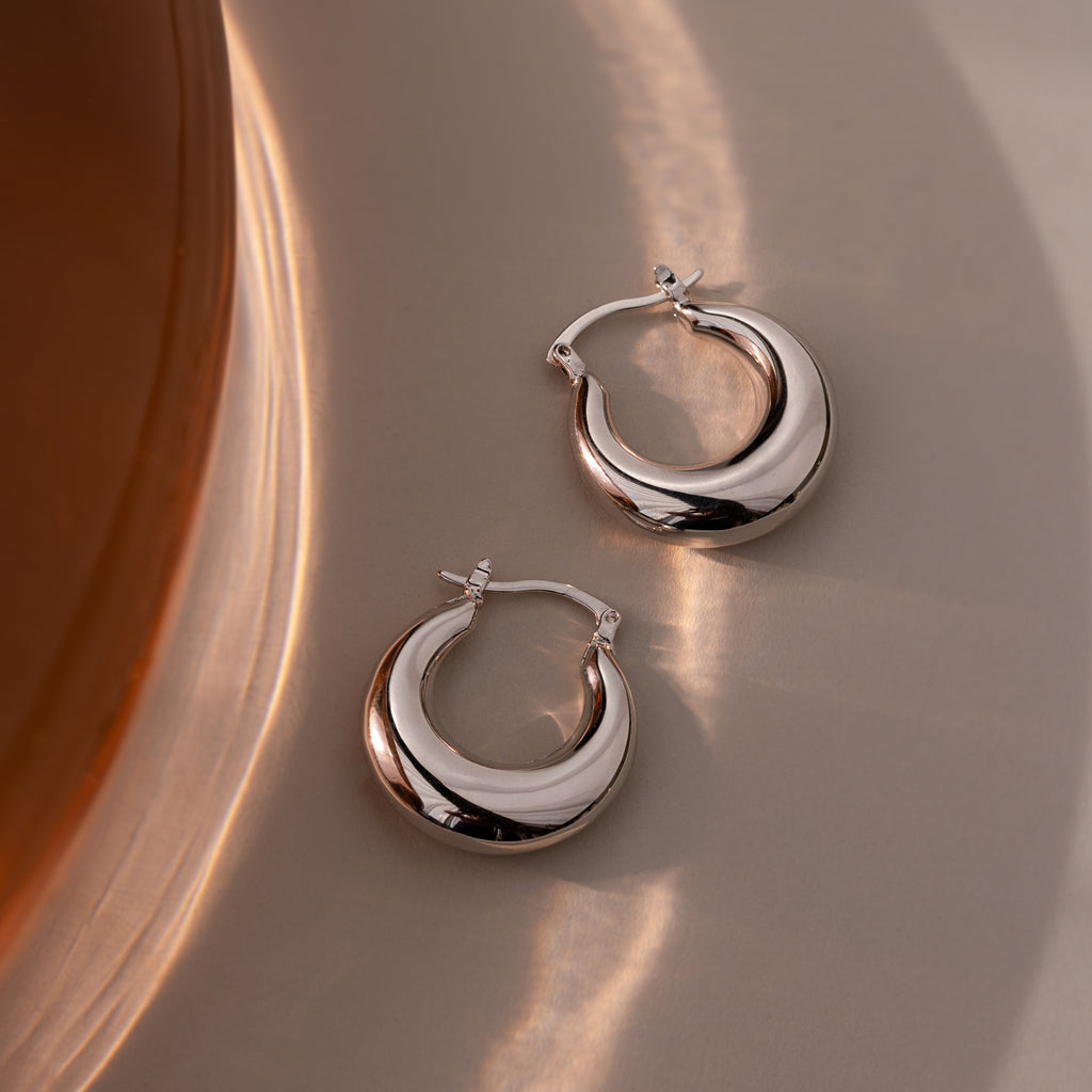 The Sculpted Hoops in White Gold feature a high-shine finish, resting on a smooth beige surface and reflecting soft light—modern classic earrings with a sleek, polished look.