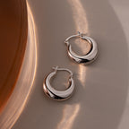 The Sculpted Hoops in White Gold feature a high-shine finish, resting on a smooth beige surface and reflecting soft light—modern classic earrings with a sleek, polished look.