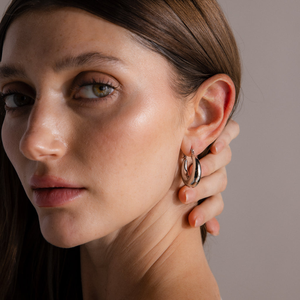A woman with straight brown hair wears Sculpted Hoops in White Gold—modern, high-shine earrings—gently holding her neck as she faces the camera.