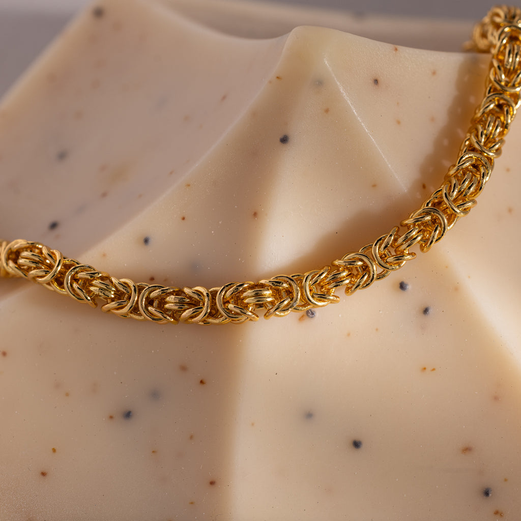 The Bold Byzantine Bracelet in 18K Gold, featuring woven links, drapes elegantly over a beige geometric object, radiating quiet luxury.