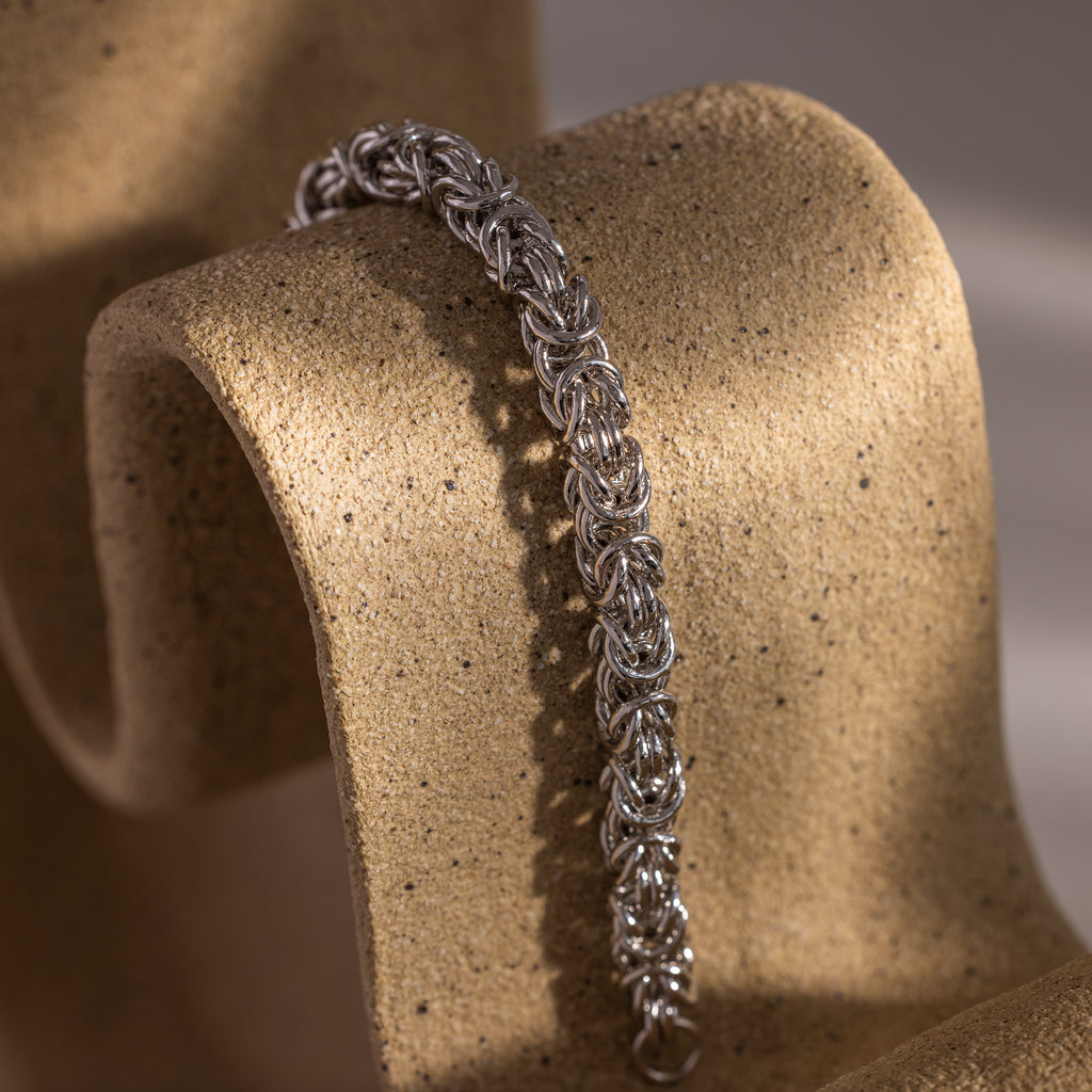 The Bold Byzantine Bracelet in white gold is showcased on a textured, curved beige surface with soft lighting, exuding the elegance of luxury jewelry.