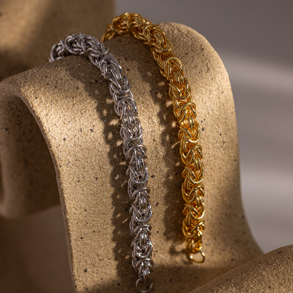 The Bold Byzantine Bracelet, featuring intricate woven links and a quiet luxury style, is displayed on a textured beige surface.