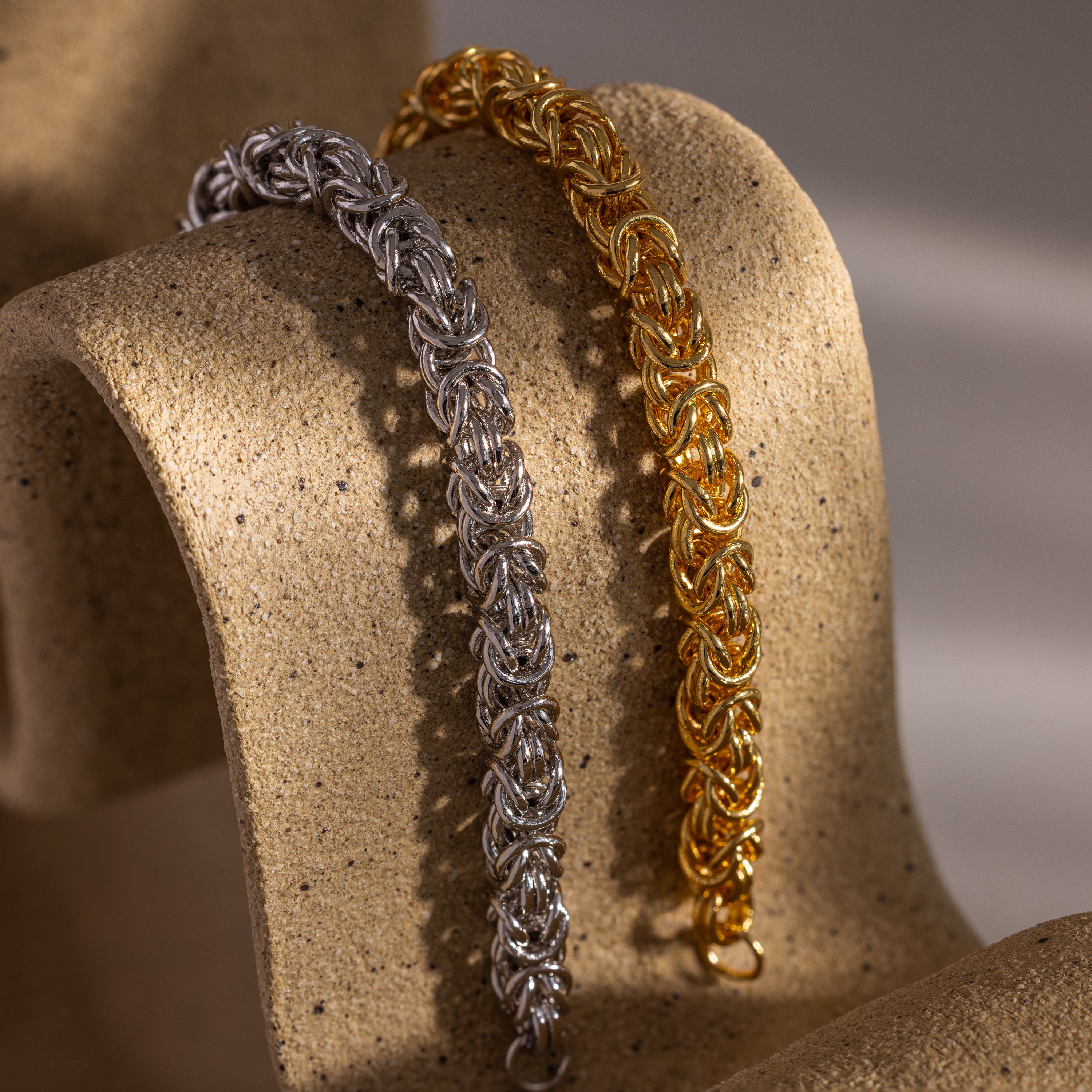 The Bold Byzantine Bracelet, featuring intricate woven links and a quiet luxury style, is displayed on a textured beige surface.