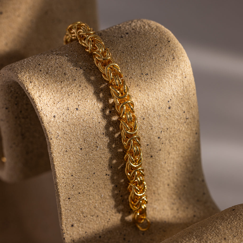 The Bold Byzantine Bracelet in 18K Gold, featuring woven links, rests elegantly on a textured, curved beige stand and embodies quiet luxury.
