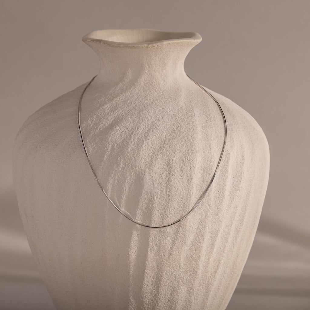 The Dainty Herringbone Necklace in Sterling Silver drapes elegantly over a textured, off-white ceramic vase for a refined layered look.